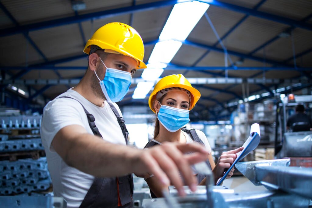 Industrial workers with face masks protected against corona virus discussing about metal parts in factory.